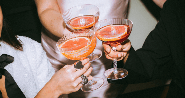 Three friends at a wedding, clinking signature cocktails garnished with an orange