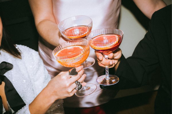 Three friends at a wedding, clinking signature cocktails garnished with an orange